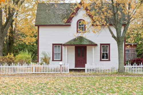 White house with red trim, red front door and picket fence out front with fall leaves on the ground