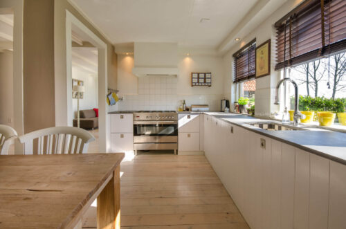 Simple kitchen layout with white cabinets, wooden floors and table and windows looking into backyard