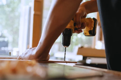 Person holding down a piece of wood while drilling into it with an electric drill