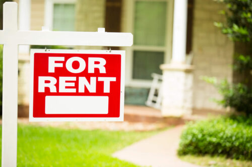 Red and white "House for Rent" sign in front of a stone, wood house that is a rental property.